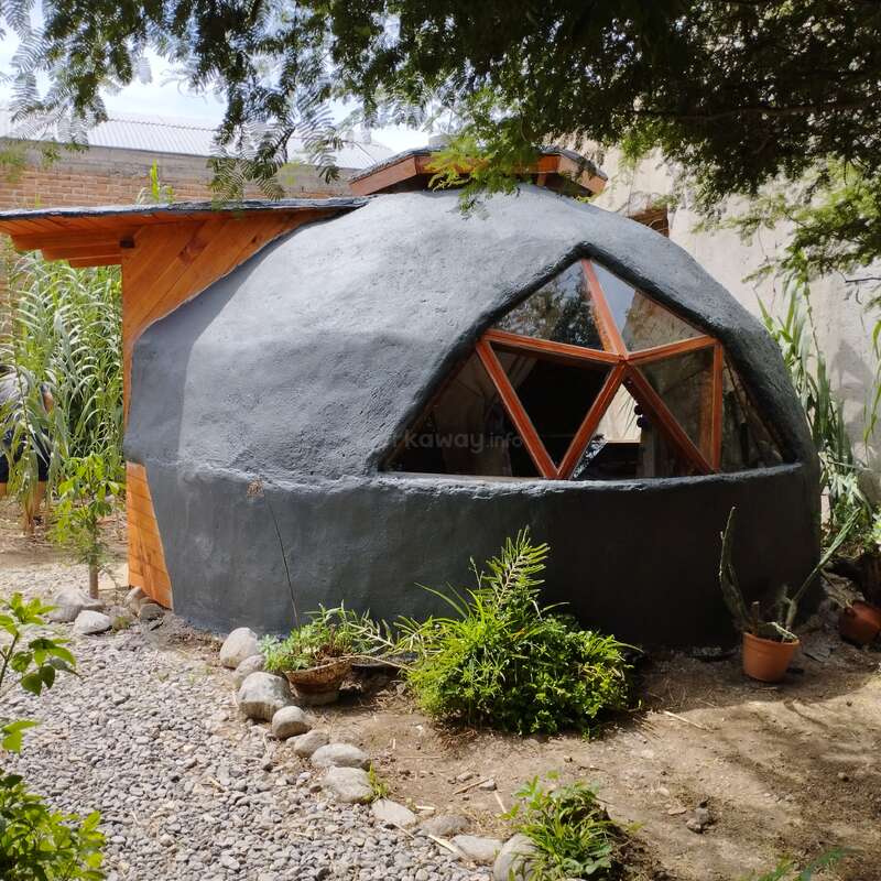 Esta imagem mostra uma pequena casa de adobe em forma de cúpula com janelas geométricas, cercada por plantas e cascalho, combinando materiais de construção naturais com um ambiente externo tranquilo.