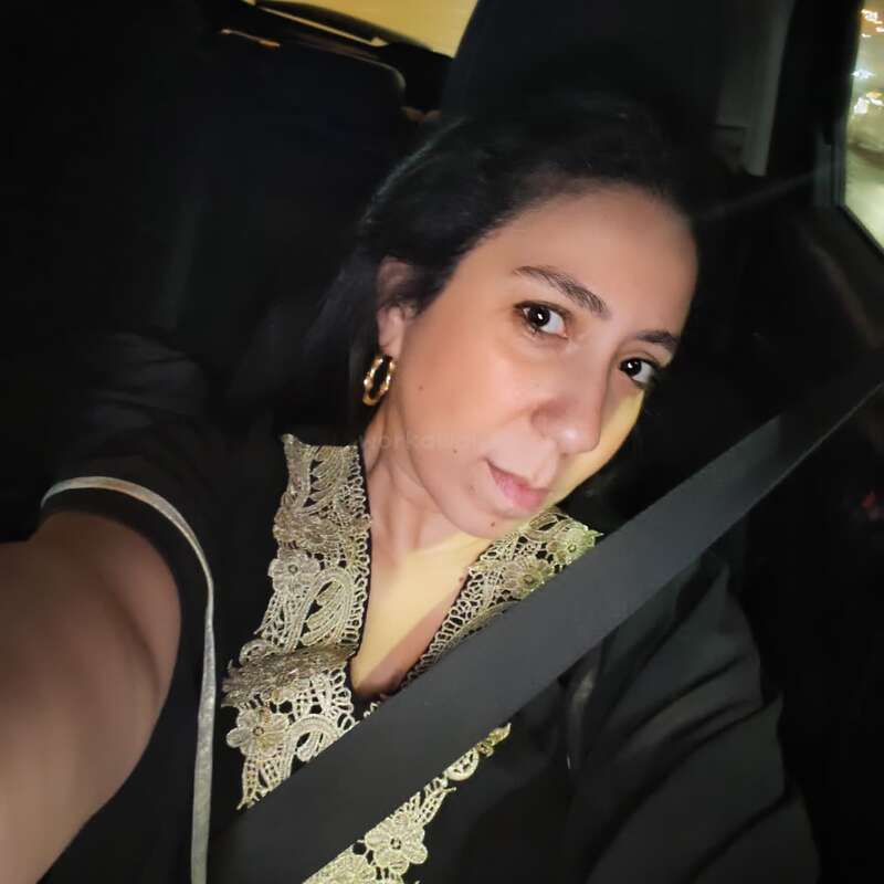 Una mujer está sentada en un coche, vestida con una prenda negra con intrincados bordados dorados. Lleva pendientes de aro dorados y el cinturón de seguridad puesto, y se está haciendo un selfi.