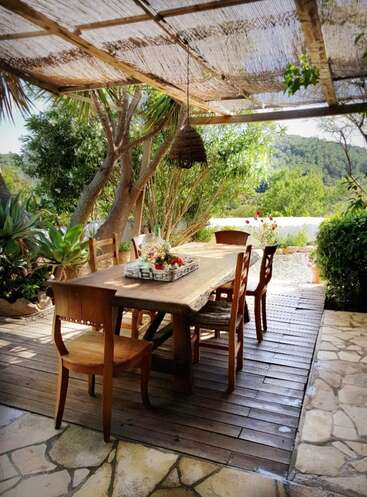 L'image montre une salle à manger extérieure sereine avec une table en bois, des chaises et un toit de chaume, entourée d'une végétation luxuriante et d'une vue pittoresque.
