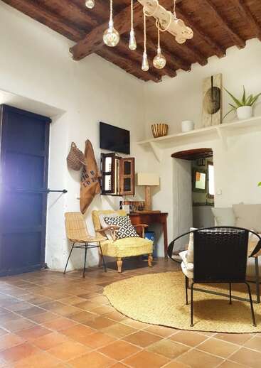 L'image représente un salon confortable aux murs blancs, au plafond en bois et aux carreaux de terre cuite, avec un coin salon confortable et des éléments décoratifs.