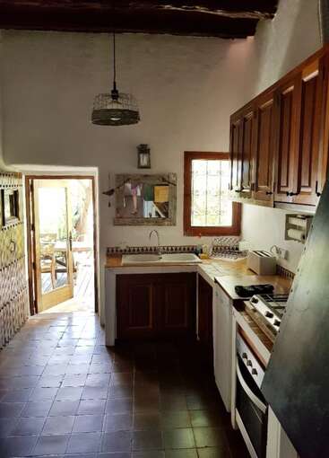 L'image représente une cuisine rustique avec des murs blancs, des armoires en bois et un sol carrelé, comprenant un évier, une cuisinière et un four, avec une porte menant à un patio extérieur.