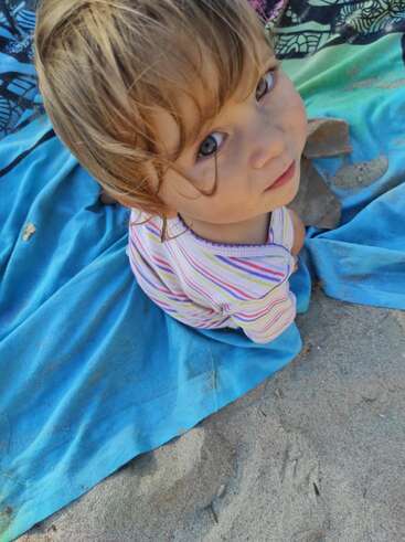 L'image représente un jeune enfant assis sur une couverture bleue, entouré de sable, avec une chemise rayée et une expression curieuse, regardant vers le haut.