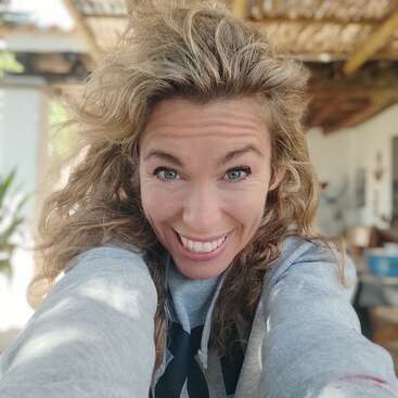 L'image représente une femme aux longs cheveux blonds ondulés, vêtue d'un sweat à capuche gris, souriant à l'appareil photo, sur un fond flou représentant une pièce aux murs blancs et au plafond en bois.