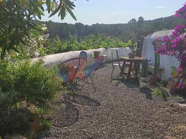 L'image représente un espace extérieur serein avec une table et des chaises, entouré de verdure luxuriante et de fleurs éclatantes, dans un paysage pittoresque.
