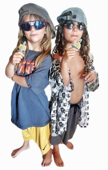 Zwei Kinder posieren selbstbewusst mit übergroßen Shirts, Sonnenbrillen und umgekehrten Mützen. Jedes von ihnen hält einen Schnuller in der Hand und drückt damit eine verspielte, coole und schrullige Haltung aus, die Spaß macht.