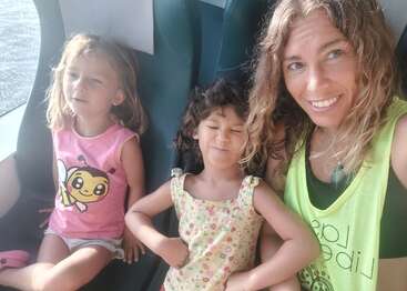 Eine lächelnde Frau und zwei junge Mädchen sitzen zusammen in einem Fahrzeug, wahrscheinlich einem Bus oder Zug. Die Stimmung ist fröhlich und hell, das Sonnenlicht scheint herein.