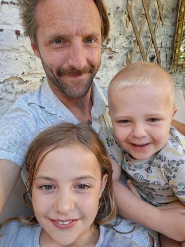 Ein Mann mit Schnurrbart lächelt, während er ein Selfie mit zwei kleinen Kindern macht, einem älteren Mädchen und einem jüngeren Jungen, die alle glücklich und fröhlich aussehen.