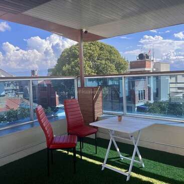 Cette image montre un balcon avec du gazon artificiel, deux chaises rouges, une table pliante blanche, des garde-corps en verre transparent et une vue sur des bâtiments et des arbres.