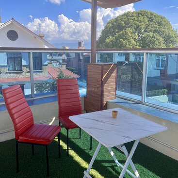 Deux chaises rouges et une petite table blanche sont posées sur du gazon artificiel. Il y a une seule tasse, une balustrade en verre, un temps ensoleillé, des arbres, des maisons et des montagnes à l'extérieur.