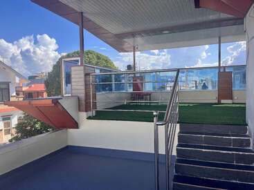 Cette image montre un toit-terrasse moderne avec gazon artificiel, toit couvert, garde-corps en verre et sièges extérieurs. Le ciel bleu et les nuages créent une atmosphère agréable et ouverte.