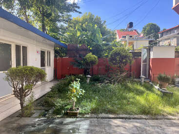 Un petit jardin avec divers arbustes et plantes est entouré d'un mur rouge. Il y a un bâtiment blanc sur la gauche, la lumière du soleil et les ombres sont présentes.