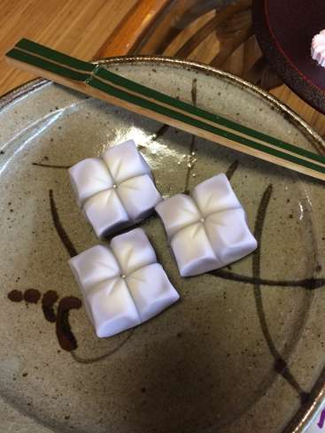 L'image représente trois bonbons blancs de forme carrée sur une assiette en céramique sur laquelle repose une baguette verte. L'assiette est ornée de motifs bruns.