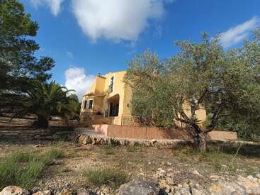 Uma villa amarela rústica fica em meio a uma paisagem seca e rochosa com árvores e palmeiras espalhadas, sob um céu azul brilhante pontilhado de nuvens brancas. Cenário tranquilo.