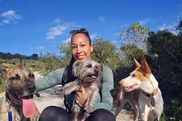 Uma mulher sorridente está sentada ao ar livre em um dia ensolarado, cercada pela natureza, segurando um pequeno cachorro fofo, com dois outros cães felizes sentados ao seu lado.