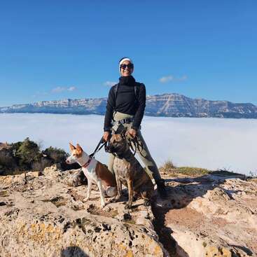 Una persona sonríe de pie sobre un acantilado rocoso con dos perros, contemplando una impresionante vista de montaña por encima de las nubes, bajo un cielo azul claro y brillante.