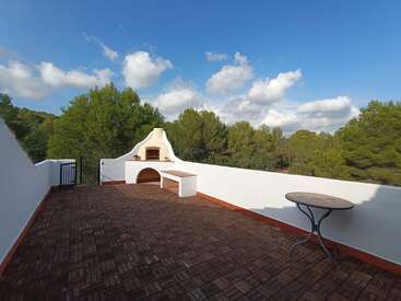 Um espaçoso terraço ao ar livre com uma churrasqueira embutida, uma mesa redonda, piso de azulejos, paredes brancas, cercado por árvores verdejantes sob um céu azul brilhante.