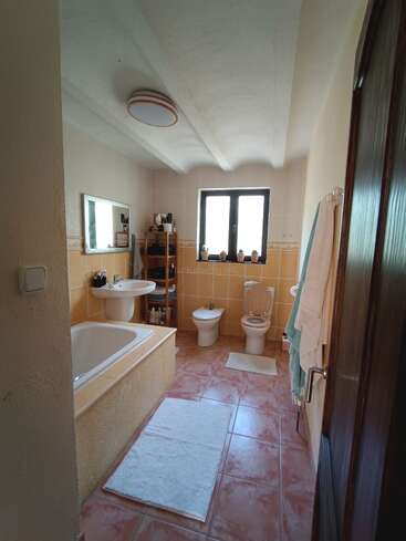 Das helle Badezimmer verfügt über eine Badewanne, ein Waschbecken, eine Toilette, ein Bidet, ein Fenster, ein Regal, einen Spiegel, Handtücher, weiße Teppiche und hellbraune Fliesen an Boden und Wänden.