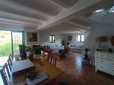 Das helle, geräumige Wohnzimmer verfügt über einen hölzernen Esstisch mit Pflanzen, bequeme Sofas, große Fenster, einen gefliesten Boden, gemütliche Sessel, Wandkunst und üppiges Grün draußen.