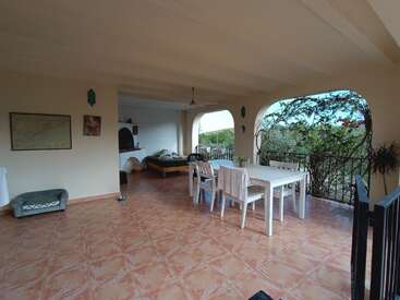 Eine geräumige, überdachte Terrasse mit gefliestem Boden, weißem Esstisch, vier Stühlen, einem Hundebett, einer Sitzecke, Wandkunst, Grünpflanzen und einem malerischen Blick ins Freie.