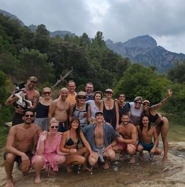 Eine Gruppe von Freunden posiert fröhlich im Freien an einem Fluss, umgeben von üppigem Grün und Bergen. Einige tragen Badekleidung und lächeln glücklich, genießen die Natur und die Zweisamkeit.