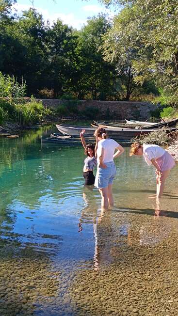 Three young women enjoy a sunny day by a clear creek, wading in shallow water, surrounded by trees and boats, relaxed and smiling together.