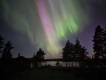 The image shows vibrant green and purple northern lights illuminating the night sky above a tranquil lake, silhouetted trees, and a dark cabin in the foreground.