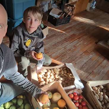 Un niño y un adulto están sentados en una habitación con cajas de frutas y verduras variadas. El niño sostiene un caqui, rodeado de patatas, manzanas y calabazas.