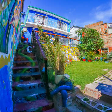 Das Bild zeigt ein lebhaftes, mehrstöckiges Gebäude mit einem Balkon, umgeben von einem üppigen Garten und einer mit bunten Graffiti verzierten Treppe, vor einem strahlend blauen Himmel.