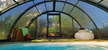 Cette image montre une piscine intérieure dotée d'une enceinte transparente en forme d'arche. Des serviettes et des flotteurs sont posés près de la piscine, tandis que la lumière du soleil filtre à travers, mettant en valeur la verdure luxuriante à l'extérieur.