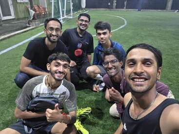 Seis jovens estão em um campo de futebol gramado, sorrindo amplamente para a câmera, sugerindo camaradagem e diversão após o jogo. A iluminação brilhante destaca seus rostos felizes e relaxados.