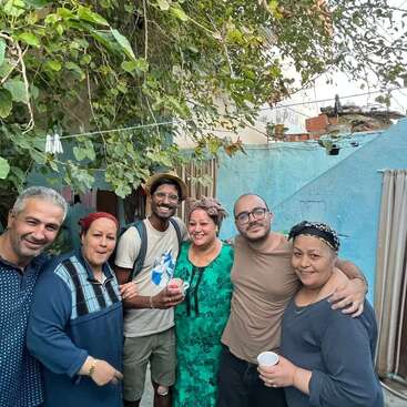Seis pessoas sorriem juntas ao ar livre, sob uma folhagem verde, contra uma parede azul. Elas parecem felizes, segurando xícaras, desfrutando de uma reunião casual em um ambiente aconchegante.