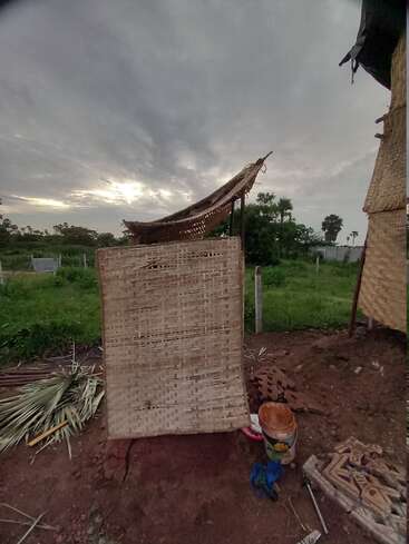 Uma pequena cabana improvisada com folhas de palmeira trançadas fica em solo descoberto, cercada por vegetação e ferramentas, sob um céu nublado ao anoitecer em uma área rural.