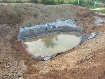 Esta imagem mostra um pequeno lago artificial parcialmente construído, forrado com plástico preto, contendo um pouco de água. A área ao redor é de solo descoberto e terreno acidentado.