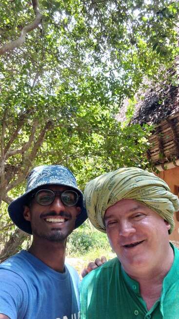 Dos hombres sonrientes posan para hacerse un selfi al aire libre. Uno lleva gafas y un sombrero de pescador, y el otro, un turbante verde. El fondo está cubierto de una exuberante vegetación.