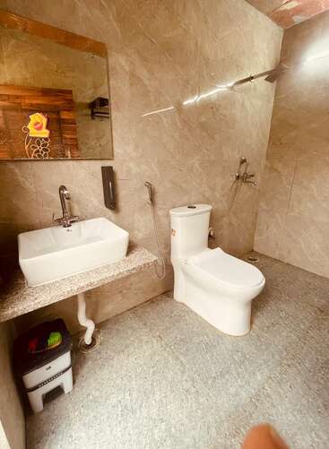 This image shows a modern bathroom with beige-tiled walls, a white sink, a mirror, a toilet, a small waste bin, and a wall-mounted faucet.