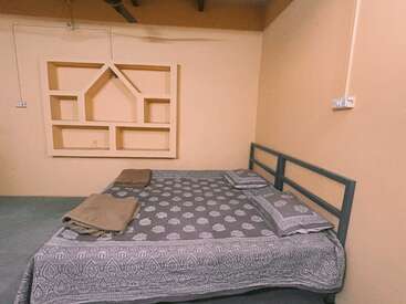 A simple bedroom features a double bed with patterned bedding, two pillows, folded blankets, peach walls, a geometric wall shelf, and minimalistic decor.