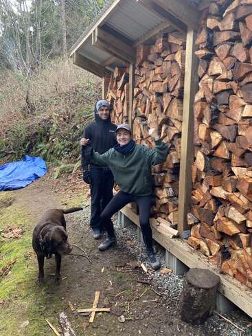 Zwei Menschen posieren fröhlich vor einem großen, ordentlich gestapelten Holzstapel unter einem Holzunterstand, während draußen in der Natur ein brauner Hund mit einem Stock daneben steht.