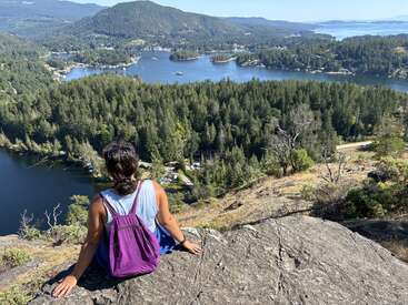 Eine Person mit einem lila Rucksack sitzt auf einem Felsvorsprung und blickt auf eine atemberaubende Waldlandschaft mit Seen, Bergen und einem strahlend blauen Himmel in der Ferne.