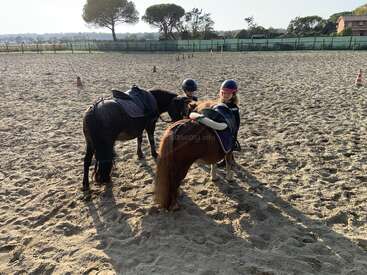 Two children wearing helmets are riding ponies in a sandy outdoor arena. They appear to be in a riding lesson on a sunny day, surrounded by cones.