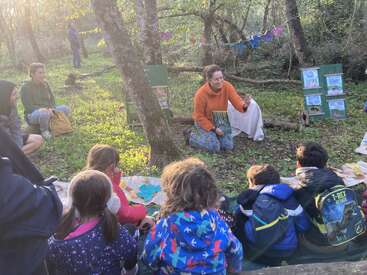 A group of children and adults sit in a sunlit forest, attentively listening to a storyteller with visual aids, surrounded by trees and colorful crafts.