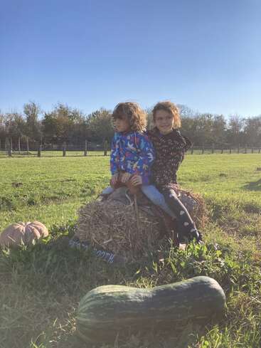 Two children sit on a hay bale in a sunny field, surrounded by pumpkins and gourds, enjoying a clear blue sky and autumn scenery.