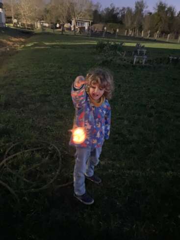A young child stands outdoors at dusk, holding a glowing object. The background features grass, scattered trees, and a small shed under a dim sky.