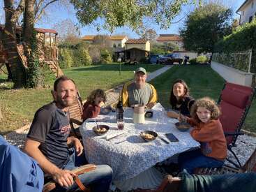 Five people enjoy an outdoor meal at a table in a sunny garden, surrounded by trees, grass, and houses, creating a warm, relaxed family atmosphere.