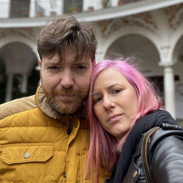 L'image représente un homme et une femme posant pour un selfie devant un bâtiment blanc à arcades, illustrant leur relation chaleureuse et affectueuse.
