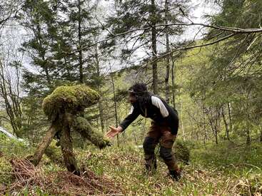 Una persona en el bosque extiende la mano hacia una formación arbórea cubierta de musgo que se asemeja a una criatura. La escena es caprichosa y combina naturaleza e imaginación.