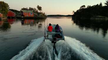 Dos personas en una lancha motora navegan a toda velocidad por aguas tranquilas al atardecer, pasando junto a coloridas cabañas rojas en una pintoresca costa arbolada rodeada de serena naturaleza.