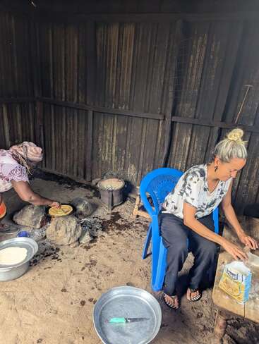 Two women are preparing and cooking food in a rustic kitchen. One rolls dough at a table, while the other cooks flatbread over an open fire.