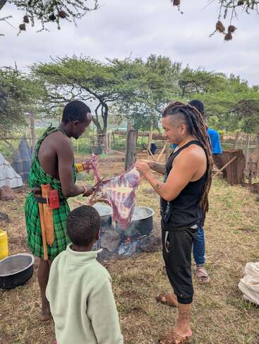 Two men and a child prepare fresh meat over an outdoor fire, surrounded by trees and rustic fences, engaging in traditional cooking in a rural setting.