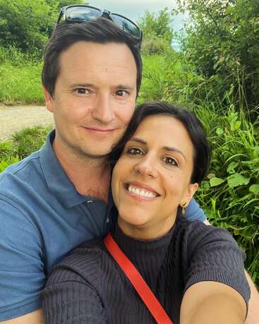 A happy couple takes a close-up selfie outdoors, smiling brightly. They are surrounded by lush green foliage, enjoying a sunny day together in nature.
