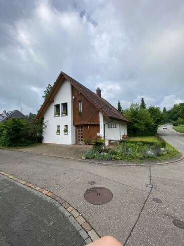 A quaint house with a steep roof stands at a street corner, surrounded by greenery and flowerbeds, under a cloudy sky on a quiet neighborhood road.